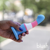 A hand with purple-painted nails holds the Avant Blush Avant P2 Silicone Pride True Blue dildo, featuring blue, white, and pink stripes. Made from body-safe silicone with a curved tip, it appears against a blurred background.